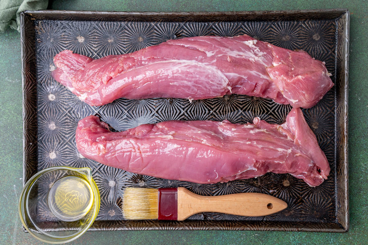 two raw pork tenderloins on a baking sheet with pastry brush and oil