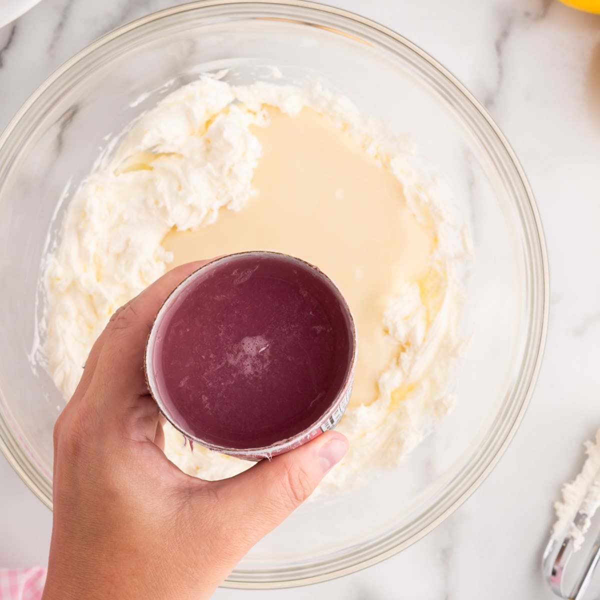 a hand holding pink lemonade concentrate over a bowl of cream cheese