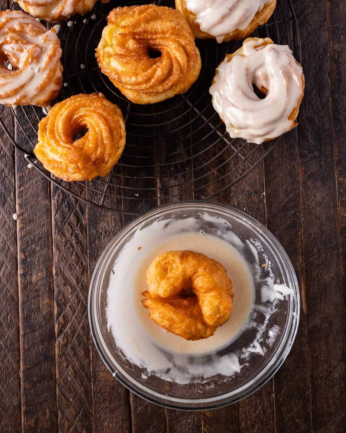 fried crullers on a cooling rack, with one dipping in glaze